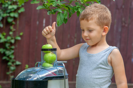 Boy child makes juice from green apples in juicer outdoors.