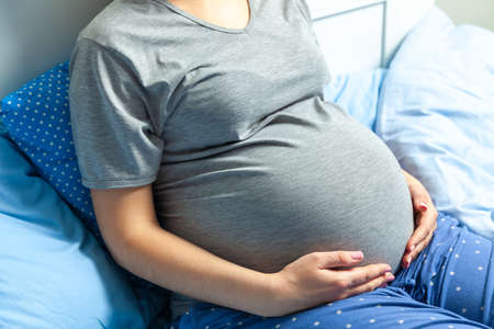Pregnant woman holding her belly and sitting on the bedの写真素材