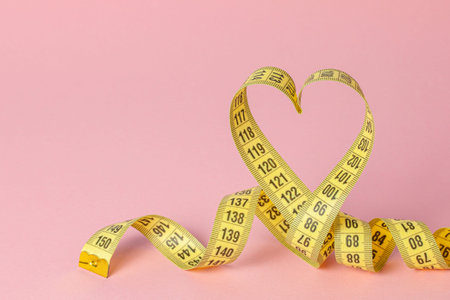 Yellow measuring tape in the shape of a heart on a pink background. The concept of weight loss for...