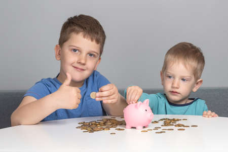Two children put coins in piggy bank. Boys are investing in their future education. The boy smiles and shows thumbs upの写真素材