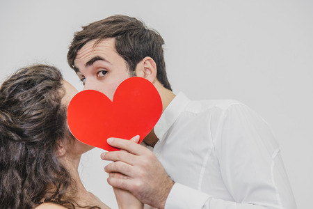 Young tender and in love couple. Hold a paper red heart in your hands. During this kiss and the man looks at the camera.の写真素材