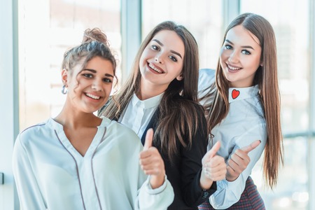 Three young business girls in the office. Dressed in classic clothing style, showing thumbs up with a gesture with copy spaceの写真素材