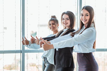 Three young business girls in the office. Dressed in classic clothing style, showing thumbs upの写真素材