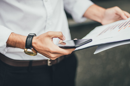Businessman using mobile phone while holding folder and documents in his hand, close up photo. Education, business, lifestyle conceptの写真素材