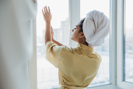 The full length of the presentation is a young woman standing by the window, enjoying the morning looking at the city after awakening in the morning. In a dressing gown with a towel on his head, resting from business, waiting for good weather.の写真素材