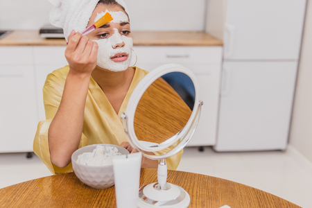 Face and body skin care, face treatment at home concept. Happy young woman applying a white mask on her face with a brush.の写真素材