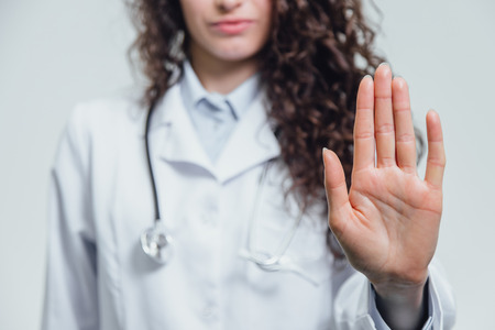 A young beautiful woman doctor gesturing stops. Probably a female doctor shows a gesture stop over a gray background.の写真素材
