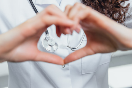 A young woman doctor is on a gray background. During this time, the heart symbol shows on the chest with his hands. Dressed in a white robe. Female doctor with a stethoscope gesturing.の写真素材