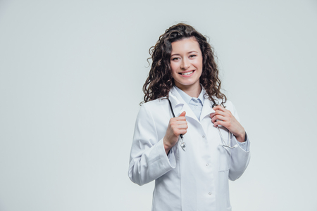 A serious young female Caucasian woman in a laboratory robe, looking at you and holding the phonendoscope out of hand. Portrait of a pretty dark hair woman - a doctor wearing white uniforms. The concept of medical treatmentの写真素材