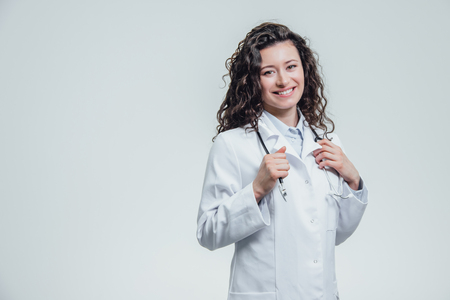 A serious young female Caucasian woman in a laboratory robe, looking at you and holding the phonendoscope out of hand. Portrait of a pretty dark hair woman - a doctor wearing white uniforms. The concept of medical treatment. Has beautiful long black curly hair.の写真素材