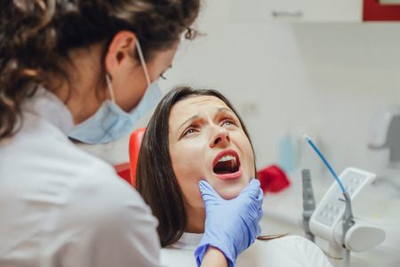 Young woman sitting in a dental chair for appointment of a doctor. During this time, she was very worried about fear, opened her big eyes. A doctor is a good woman in medical clothes, which makes a doctor.の写真素材