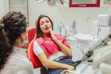 Close-up portrait of a sad young girl with a painful tooth, a doctor in office chairs, an isolated dentist office background clinic. Eの写真素材