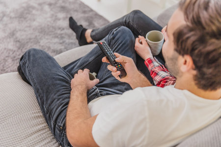 Photo of young happy happy couple sitting on a couch indoors and watching TV. Looking sideways The wife holds a cup in her hands, a mans console.の写真素材