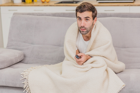 A man with a cold sitting on the couch in a blanket is as cold as possible. Man at home - sick man. Gray sofa in the room. The concept of a misfortune. The concept of health and healthy lifestyle.の写真素材