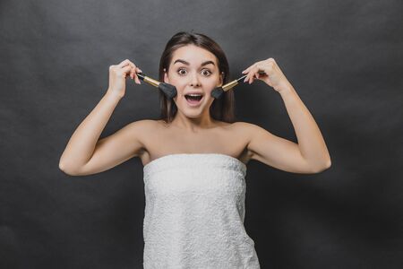 Astonished cute brunette girl in white towel with make up brushes. Isolated on black background.の写真素材