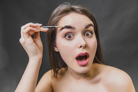 Beautiful young girl stands on a black background. During this, looking into the camera using a brush, makes an eyebrow makeup.の写真素材