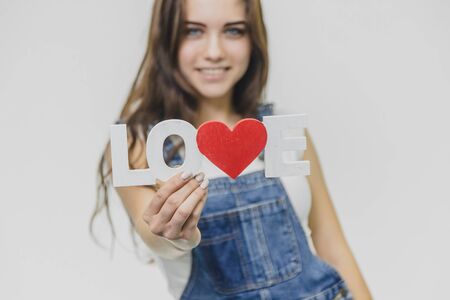 A young beautiful girl is on a gray background. Dressed in a denim suit and a white T-shirt. Holds a sign with a word of love. Opening his blue eyes smiles genuinely.の写真素材