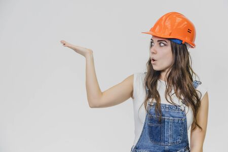 A young beautiful girl stands on a gray background. It is dressed in an orange helmet on the head. During this, he has a surprised look and looks aside. He raised his hand up.の写真素材