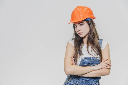 Young pretty girl standing on a white background. During this time wearing a white T-shirt and denim overalls. Dressed in an orange helmet.の写真素材