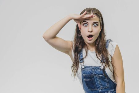 A young beautiful girl stands on a gray background. It is dressed in a denim suit and a white T-shirt. During this time she looks at the camera.の写真素材