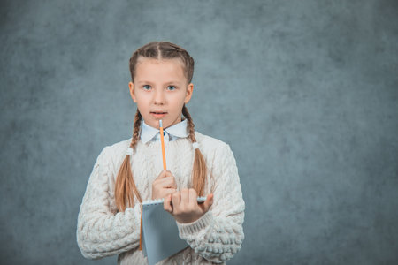 A schoolgirl in light grey sweater with braids and pencils in her hand is isolated on grey...