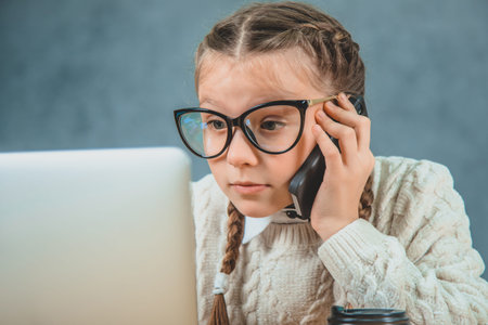 Little blond female student is on the phone and looking at the laptop with all her seriousness.