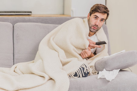 A man with a cold sitting on a sofa in a light blanket. Man at home is a young man. Gray sofa in the...