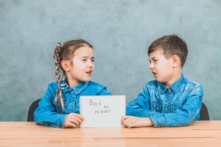 Pretty boy and girl sitting at the desk holding a sheet of paper with the phrase back to school written on it. They dont like the idea of returning to school.の写真素材