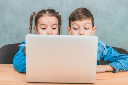 Cute little schoolkids sitting at the desk, surprised by something they have seen in the laptop.