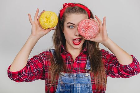 Childish young girl is hiding her eye behind big doughnut with pink icing, holding yellow doughnut near head, mouth widely open.の写真素材