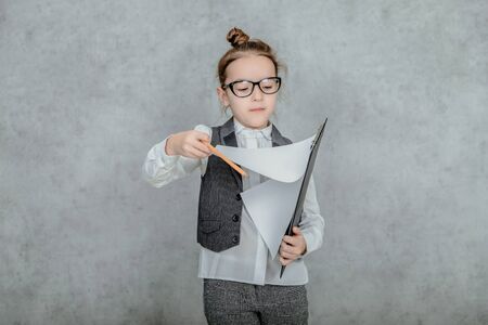 Little girl with a folder in her hands on a gray background. Dressed up as a business lady and glasses. Viewing business documents.の写真素材