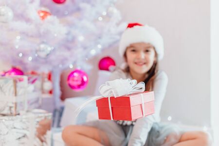 Blurred smiling santa child sitting under fir-tree, extending a gift wrapped for Christmas to the camera. Focus on the red box.の写真素材