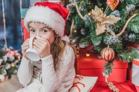 Close-up photo of cute smiling girl sitting cozy at home, drinking hot chocolate at christmas tree on wooden background.の写真素材