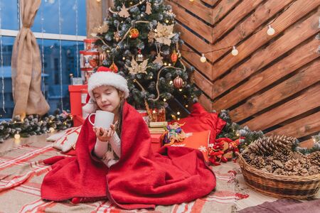 Close-up photo of cute smiling girl sitting cozy at home, drinking hot chocolate at christmas tree on wooden background.の写真素材
