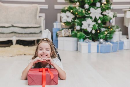 Closeup of beautiful little angel girl with perfect smile laying on the floor, keepin hands on big present box with ribbon.の写真素材