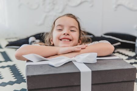 Closeup of beautiful little angel girl with perfect smile laying on the floor, keepin hands on big present box with ribbon.の写真素材