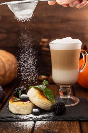 Two cheesecakes and a glass mug with latte stand on a wooden table on a black stand. Powdered sugar is poured on top of the cheesecakesの写真素材