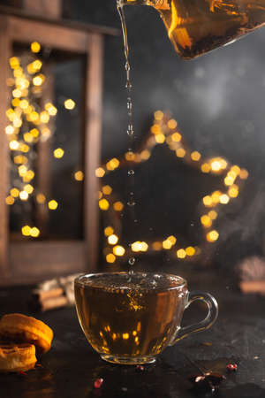 Tea from a glass teapot is poured into a glass mug on a dark background with lightsの写真素材