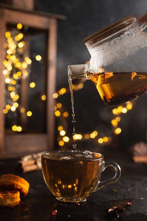 Tea from a glass teapot is poured into a glass mug on a dark background with lightsの写真素材