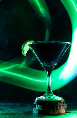a cocktail glass with a drink inside stands on a stand with a slice of lime on the edge. neon trail behind the shot glassの写真素材