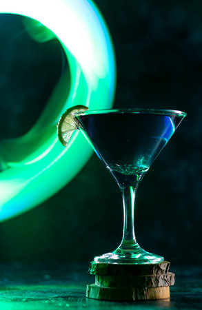 a cocktail glass with a drink inside stands on a stand with a slice of lime on the edge. neon trail behind the shot glassの写真素材