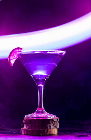 a cocktail glass with a drink inside stands on a stand with a slice of lime on the edge. neon trail behind the shot glassの写真素材