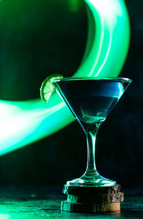a cocktail glass with a drink inside stands on a stand with a slice of lime on the edge. neon trail behind the shot glassの写真素材