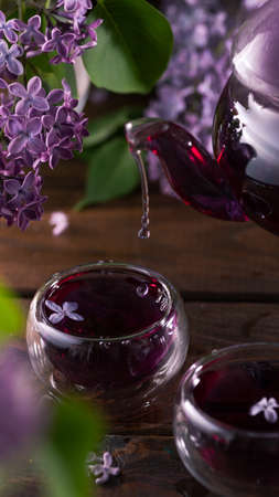 Purple tea is poured from a glass teapot into cups. Lilac flowers are all aroundの写真素材