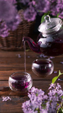 Purple tea is poured from a glass teapot into cups. Lilac flowers are all aroundの写真素材
