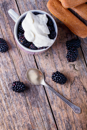 Cup of fresh natural blackberries with whipped cream and biscuits. Spoon and wooden table.の写真素材