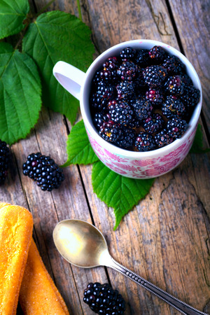 Cup of fresh natural blackberries with biscuits. Blackberries leaf. Spoon and wooden table.の写真素材