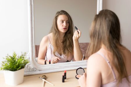 Young woman portrait putting on make up in front of a mirror with make up tools on the table. Natural light and plant.の写真素材