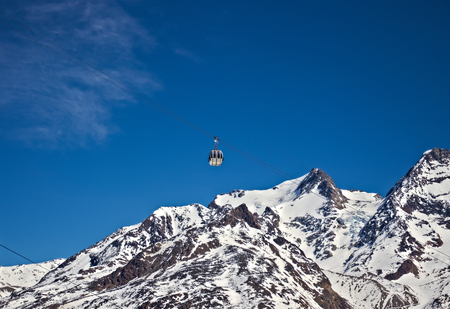 Gondola passing in front of alpine snow mountains and blue sky.の写真素材