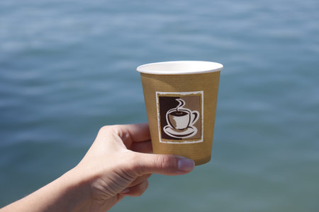 Malta, may 2019: Womens hand holding papercup with coffee in front of ocean waves. Showing recycling...